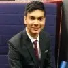 Young man in a dark suit and tie sitting on a blue couch, smiling at the camera.