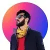 Bearded man in glasses, black jacket, yellow scarf, and red shirt, looking to the side with a colorful circle background.