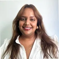 Smiling woman with long brown hair wearing a white blouse against a plain light background.