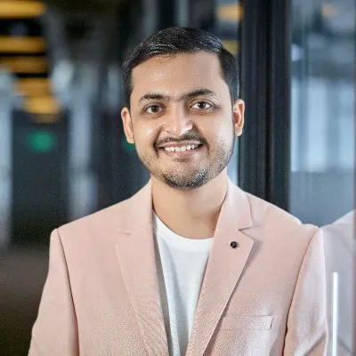 Smiling man with short dark hair wearing a light pink blazer and white shirt in a modern indoor setting.